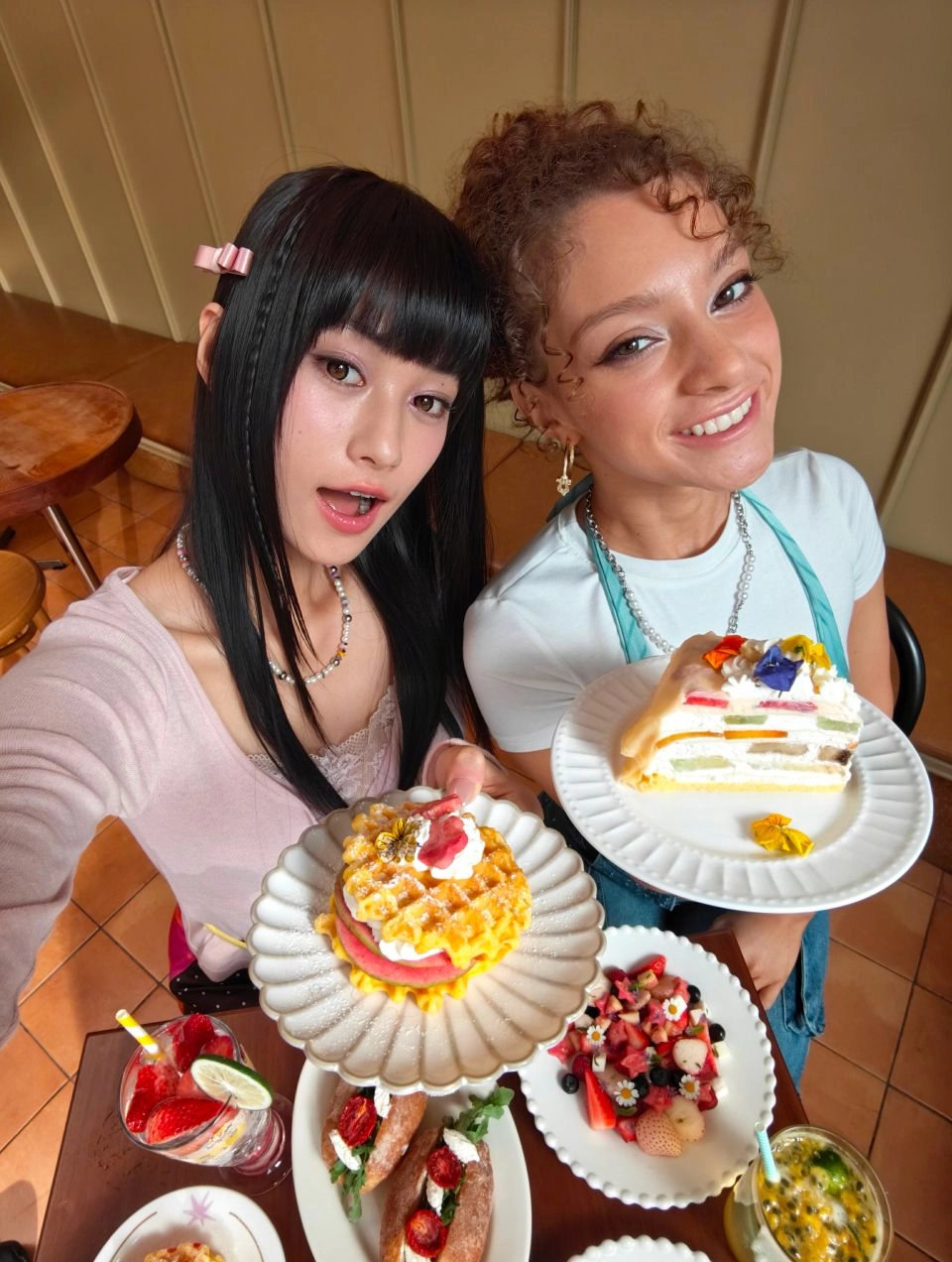 A 0.6x ultra-wide cafe selfie of two women and a table full of desserts