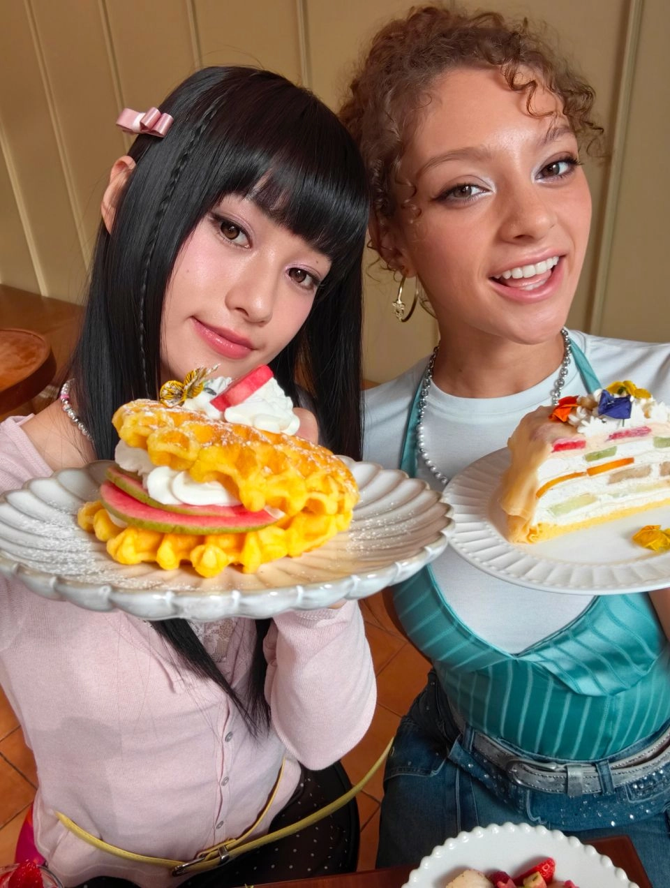 1x selfie camera sample. Two girls pose playfully with a fruit waffle and crepe cake, capturing clear facial and food details