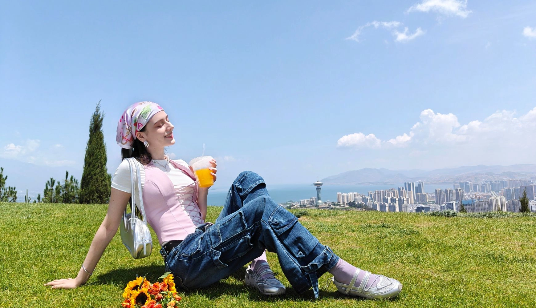 A 1x zoom photo, showing a woman sitting on grass with a sweeping city and lake background